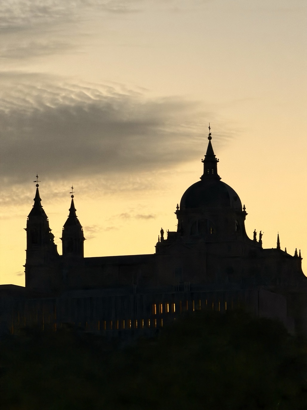 The domed silhouette of the Almudena cathedral, with its lantern spire and twin towers, rises against a golden sunset sky above a row of small glowing windows.