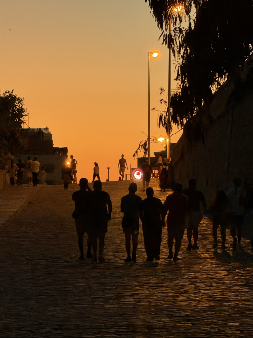 People walk in silhouette up a cobbled street toward a glowing orange sunset, lit street lamps lining a high wall on the right.