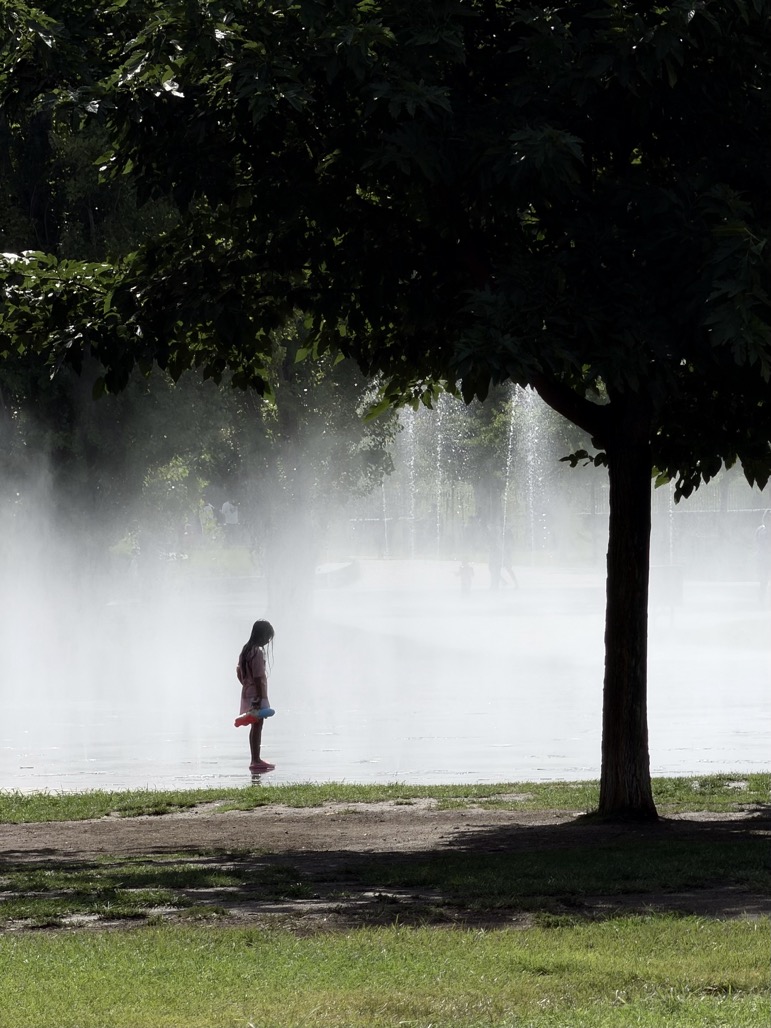 A young girl stands alone at the edge of a misty splash fountain, water jets spraying behind her through the haze, framed by a large shady tree on a summer day.
