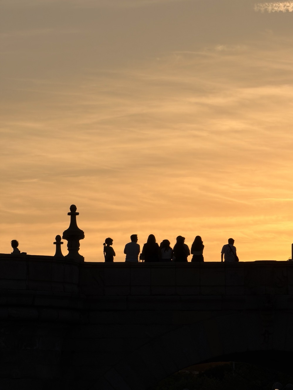 A group of people stand silhouetted along the parapet of an old stone bridge against a glowing orange sunset sky streaked with thin clouds.