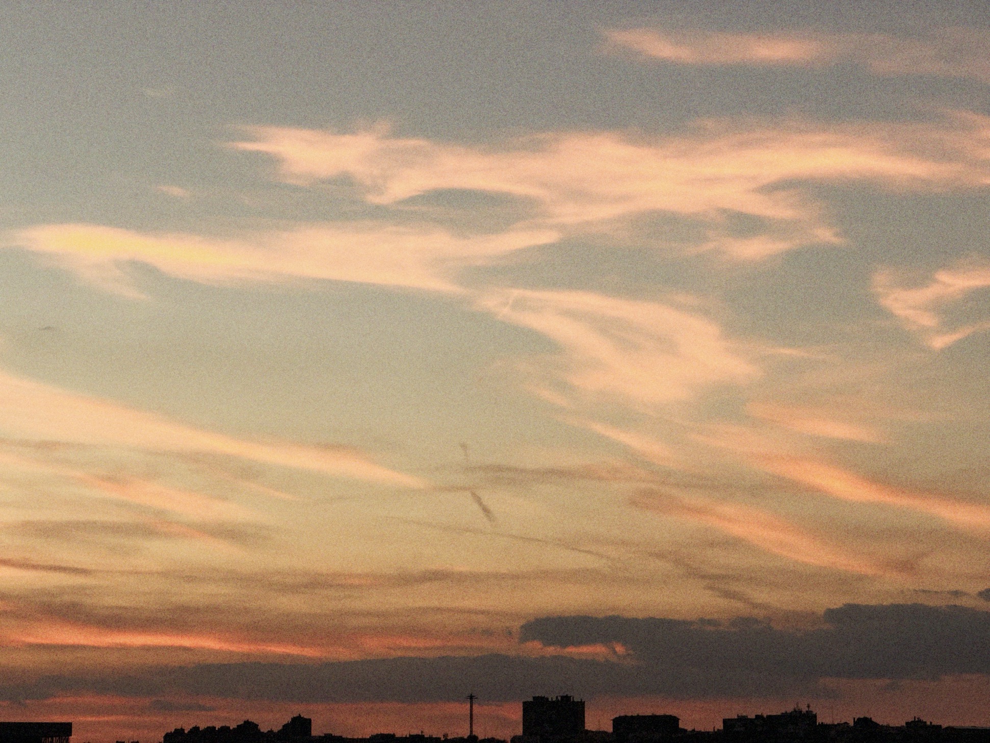 Sweeping wisps of cloud tinted pink and orange fill a vast sunset sky above a thin strip of silhouetted city rooftops.