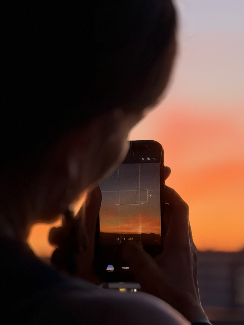 A person seen from behind holds a smartphone to capture a vivid orange and pink sunset above dark rooftops.