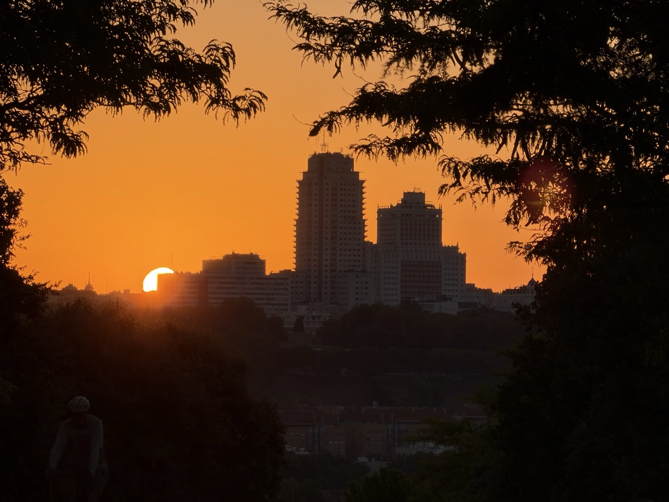 The towers of Plaza de España stand silhouetted against a glowing orange sky with the sun low on the horizon, framed by dark tree branches, a lone cyclist in the shadowed foreground.