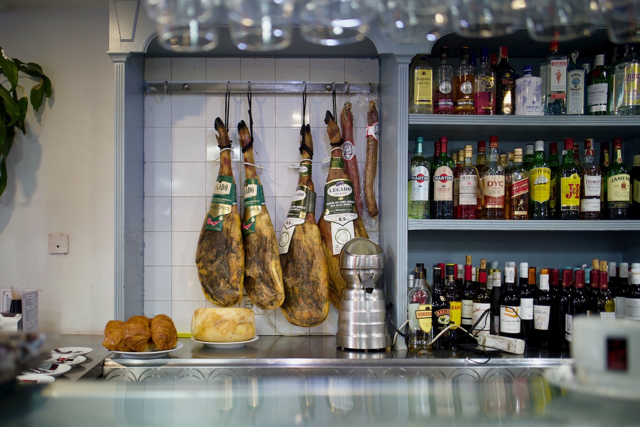 Hams hanging, bottles on shelves, bread and pastries on counter in a bar or restaurant..
